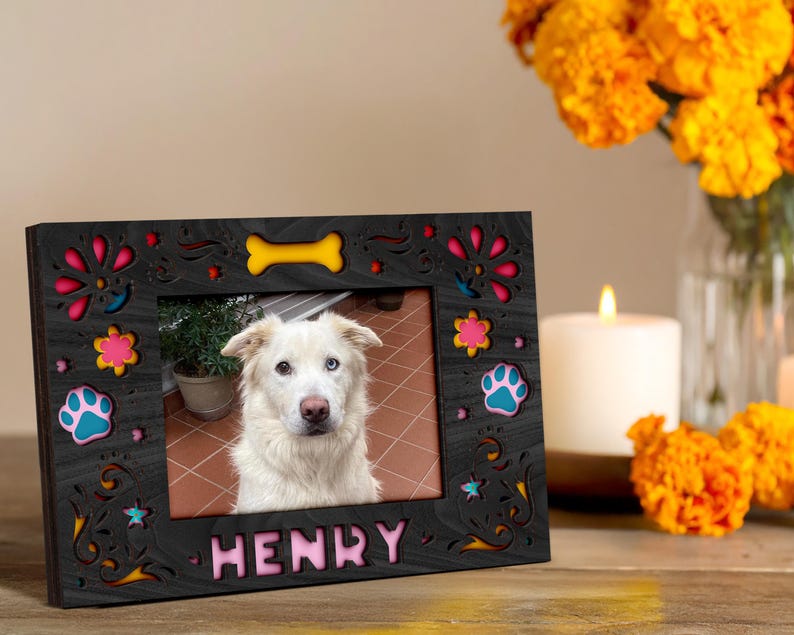 May include: A black wooden picture frame with colorful floral and paw print cutouts. The frame holds a photo of a white dog with the name "HENRY" in pink letters. A yellow bone cutout is at the top.