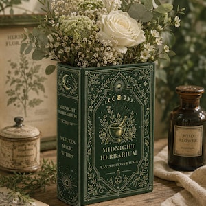 May include: A dark green rectangular vase shaped like a book titled Midnight Herbarium, featuring gold celestial and botanical illustrations, filled with a bouquet of white roses, baby's breath, and eucalyptus leaves.