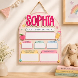 May include: A wooden arch-shaped milestone board featuring the name SOPHIA in pink letters at the top. The board includes text fields for date, age, height, future career, and interests, decorated with a pink apple, a yellow pencil, and stars.