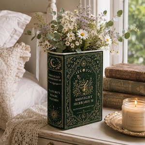 May include: A dark green rectangular vase shaped like a book titled Midnight Herbarium, featuring gold celestial and botanical illustrations. It is filled with a bouquet of white daisies, purple wildflowers, and green eucalyptus leaves.