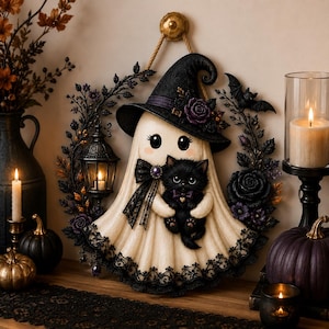 May include: A decorative wall hanging featuring a cream-colored ghost wearing a black witch hat and holding a small black kitten. The ghost is surrounded by a wreath of black roses, leaves, a small lantern, and a bat, all suspended by a rope.