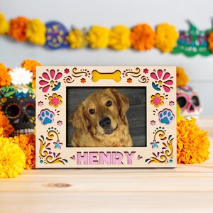 May include: Wooden photo frame with a picture of a golden retriever dog named Henry. The frame has a decorative design with pink, yellow, and blue accents. A bone-shaped cutout is at the top. The background includes marigold flowers and a Day of the Dead skull.