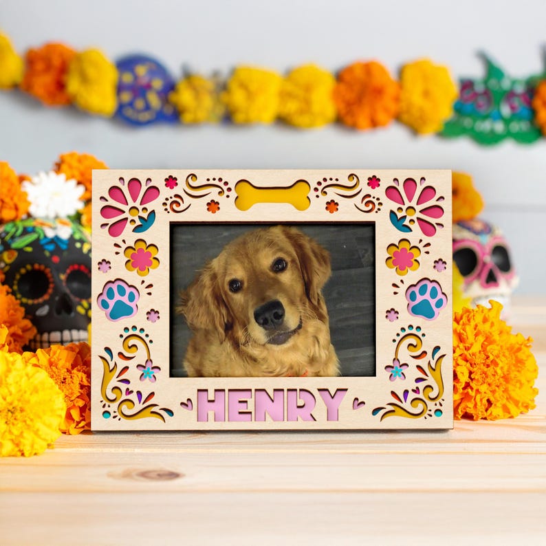 May include: Wooden photo frame with a picture of a golden retriever dog named Henry. The frame has a decorative design with pink, yellow, and blue accents. A bone-shaped cutout is at the top. The background includes marigold flowers and a Day of the Dead skull.