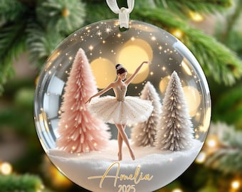 May include: A clear, round Christmas ornament featuring a ballerina in a tutu, standing in a snowy scene with pink and white trees. The ornament has the name "Amelia" and the year "2025" in gold script.