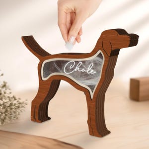 May include: A wooden dog-shaped keepsake box with a clear center filled with white material. The dog is brown with darker brown accents and the name "Chale" is written in cursive on the clear center. A hand is placing a small white item into the box.