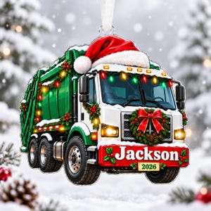 Puede incluir: Un adorno navideño con forma de camión de basura verde y blanco que lleva un gorro de Papá Noel en el techo, luces, una corona en la parrilla frontal y un parachoques rojo con el nombre Jackson y el año 2026 en blanco.