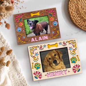 May include: Two wooden picture frames with dog photos. The frames have colorful floral and paw print designs, with the names "ALAIN" and "HENRY" in pink letters. One frame is brown, the other is light wood.