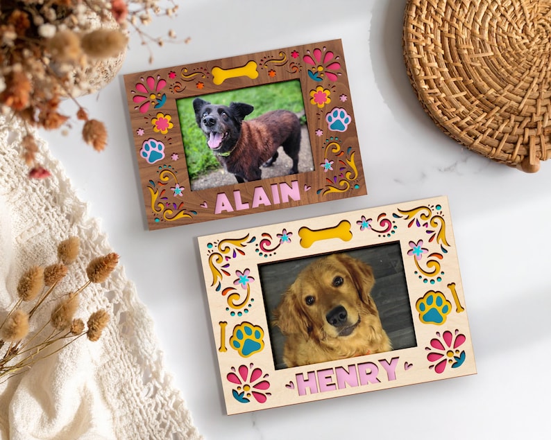 May include: Two wooden picture frames with dog photos. The frames have colorful floral and paw print designs, with the names "ALAIN" and "HENRY" in pink letters. One frame is brown, the other is light wood.