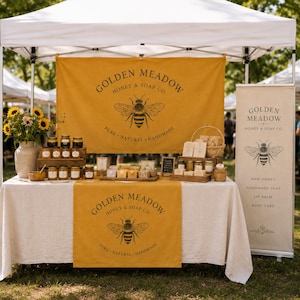 Peut inclure: Un stand de marché pour Golden Meadow Honey & Soap Co. avec une bannière jaune moutarde illustrée d'une abeille, des pots de miel, des savons, un bouquet de tournesols et une enseigne verticale listant miel brut, savon artisanal et baume à lèvres.