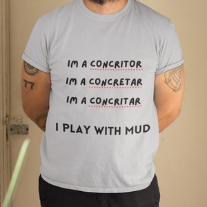 May include: A man wearing a grey t-shirt with the text "IM A CONCRITOR IM A CONCRETAR IM A CONCRITAR I PLAY WITH MUD" printed on it. He is wearing a black beanie and black pants.
