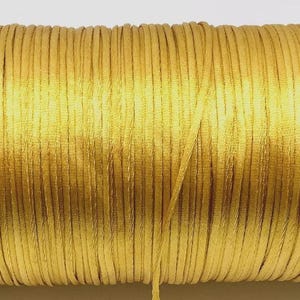 May include: A spool of golden-yellow satin cord. The cord is tightly wound around a cardboard core, with a length of cord extending from the spool. The cord is smooth and shiny, ideal for crafting and jewelry making.