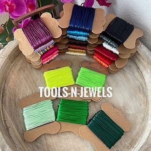 May include: A collection of colorful thread spools in various shades, including purple, blue, black, red, green, and yellow. The spools are arranged on a wooden tray, with the text "TOOLS-N-JEWELS" visible. Orchids are in the background.