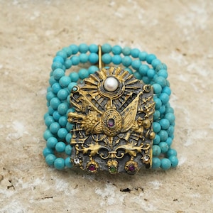 May include: A turquoise beaded bracelet with a gold and silver ornate charm featuring a sun, a pearl, and a bird.