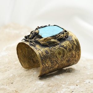 May include: A gold cuff bracelet with an ornate design and a turquoise stone set in the center. The bracelet is decorated with leaves and a small red stone.