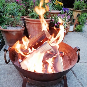 May include: A rustic fire pit with a metal bowl and three legs. The bowl is filled with burning wood and flames. The fire pit is sitting on a grey concrete surface.