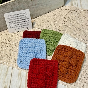 May include: Six crocheted prayer squares in different colors, including red, blue, green, white, and brown. Each square features a raised cross design. The squares are arranged on a crocheted doily. A card with the text "This pocket prayer square was made especially for you. Slip it into your pocket & through out the day touch the Cross inside and be mindful of God's love and grace for you. Keep it as a tangible symbol of God's peace +" is visible in the background.