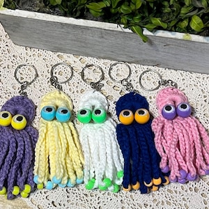 May include: Five handmade octopus keychains in purple, yellow, white, navy blue, and pink. Each keychain features large googly eyes and dangling tentacles. The keychains are made of yarn and have metal key rings.