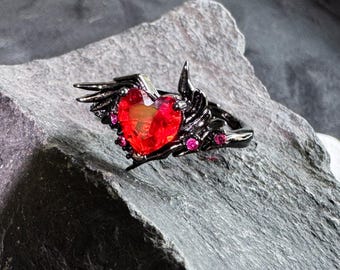 May include: A black ring with a red heart-shaped gemstone and small pink gemstones. The ring has a wing design on each side of the heart. The ring is set on a gray stone surface.