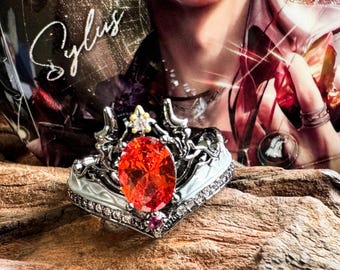 May include: A silver ring with an oval, orange gemstone at the center, surrounded by smaller clear stones. The ring has a crown-like design with a small yellow stone at the top. The ring is displayed on a piece of wood.