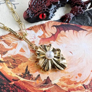May include: A gold-colored necklace with a pendant featuring a pearl center and a sunburst design. The necklace is on a patterned background with a red and black dragon figurine.