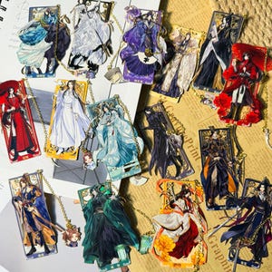 May include: A selection of acrylic charms depicting anime-style characters in diverse attire. Each charm is rectangular, with a gold chain and a small character pendant. The characters are dressed in vivid colours, including red, blue, and green.