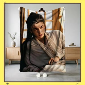May include: A beige blanket with a portrait of a person with long black hair, wearing a light-coloured robe. The person is positioned in front of a window with sunlight streaming through. The blanket is held up by a person wearing white shoes.