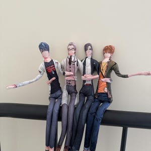 Puede incluir: Cuatro muñecos de tela hechos a mano de personajes de estilo anime, cada uno con atuendos únicos. Los muñecos están sentados en una barra negra, con los brazos extendidos. Los personajes visten atuendos que incluyen una chaqueta blanca, un chaleco y una chaqueta verde.