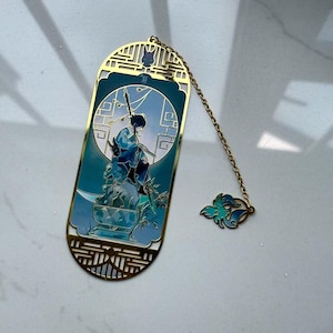 May include: A gold-colored metal bookmark with a detailed illustration of a figure in blue robes. The bookmark has a chain with a teal and blue charm. The design features intricate cut-out patterns.