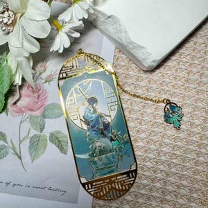 May include: A gold-colored metal bookmark with a detailed illustration of a person in blue robes, holding a weapon. The bookmark has a decorative chain with a teal and blue charm. The design is set against a light blue background.