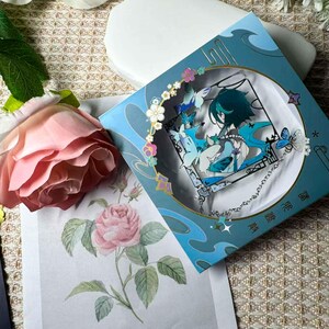 May include: A teal-colored box with a framed anime character design, surrounded by floral and butterfly accents. The box is next to a pink rose and a floral print. The design features a character with teal hair and blue accents.