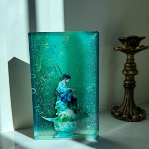 Peut inclure: Une pi&egrave;ce d'art en r&eacute;sine rectangulaire avec un fond turquoise repr&eacute;sentant une figure assise en robes bleues, des tiges de bambou et une base d&eacute;corative. Un bougeoir dor&eacute; est &agrave; droite.