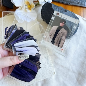 May include: A collection of clear acrylic card holders, some containing images of clothing and others with a photo of a person. The card holders are arranged in a fan shape, with a few standing upright. The background includes white flowers and a dark rock.