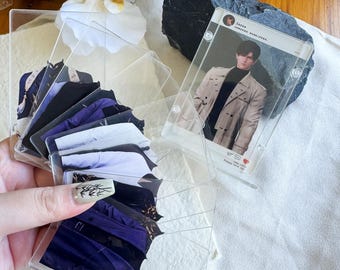 May include: A collection of clear acrylic card holders, some containing images of clothing and others with a photo of a person. The card holders are arranged in a fan shape, with a few standing upright. The background includes white flowers and a dark rock.