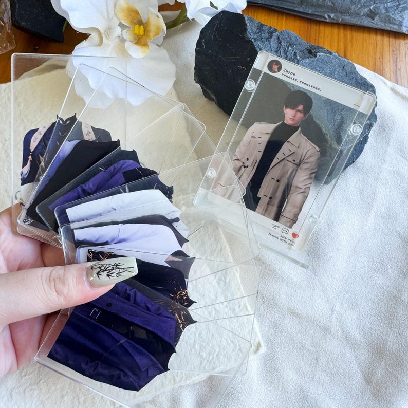May include: A collection of clear acrylic card holders, some containing images of clothing and others with a photo of a person. The card holders are arranged in a fan shape, with a few standing upright. The background includes white flowers and a dark rock.