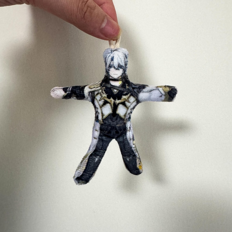 Op de afbeelding: Een kleine, handgemaakte pop in de vorm van een personage met wit haar en een gedetailleerde outfit in zwart, wit en goud. De pop heeft uitgestrekte armen en wordt aan de bovenkant opgehangen met een touwtje, wat suggereert dat het een ornament of decoratie kan zijn.