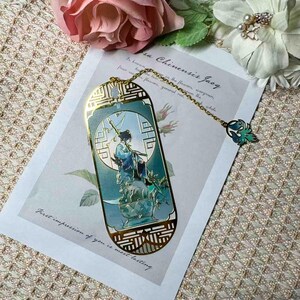May include: A decorative bookmark with a gold-colored frame and a detailed illustration of a person in traditional attire. The bookmark has a chain with a green and gold charm. The text "First impression of you is most lasting" is visible.