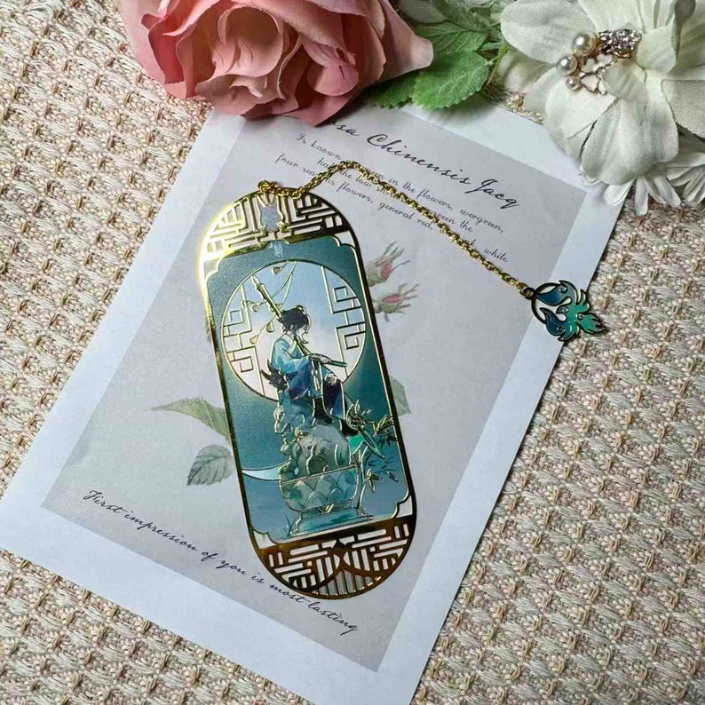 May include: A decorative bookmark with a gold-colored frame and a detailed illustration of a person in traditional attire. The bookmark has a chain with a green and gold charm. The text "First impression of you is most lasting" is visible.