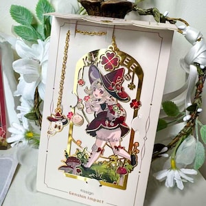 May include: A gold-colored metal bookmark featuring a cartoon character in a burgundy dress and hat, holding flowers. The bookmark has a chain with a charm and the text "Klee". The bottom of the bookmark has the text "eissign - Genshin Impact".