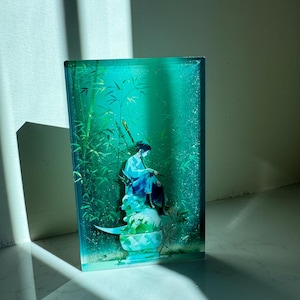 Peut inclure: Une &oelig;uvre d'art rectangulaire, de couleur sarcelle, repr&eacute;sentant une personne en robe bleue assise sur un socle d&eacute;coratif, entour&eacute;e de bambous. L'&oelig;uvre a une finition brillante et se d&eacute;tache sur un fond clair.