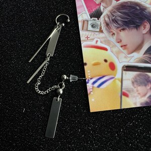 Op de afbeelding: Een paar zilverkleurige oorbellen. E&eacute;n oorbel heeft een ketting en een rechthoekige hanger, de andere een pin met een kleine bal en een rechthoekige hanger. De oorbellen liggen op een zwart oppervlak.