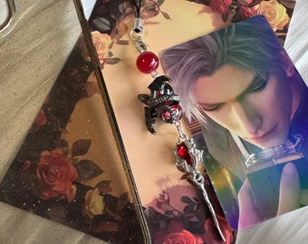 May include: A phone charm with a black cord, a red bead, and a decorative pendant featuring a dragon and a red gem. The charm is displayed with a phone case and a card with a man's portrait, all on a floral background.