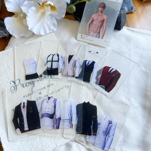 May include: A collection of clear acrylic cards featuring various suit designs, including blazers, vests, and shirts. A card with a male figure is also present. The cards are arranged on a white surface with floral accents.
