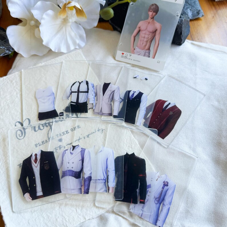 May include: A collection of clear acrylic cards featuring various suit designs, including blazers, vests, and shirts. A card with a male figure is also present. The cards are arranged on a white surface with floral accents.