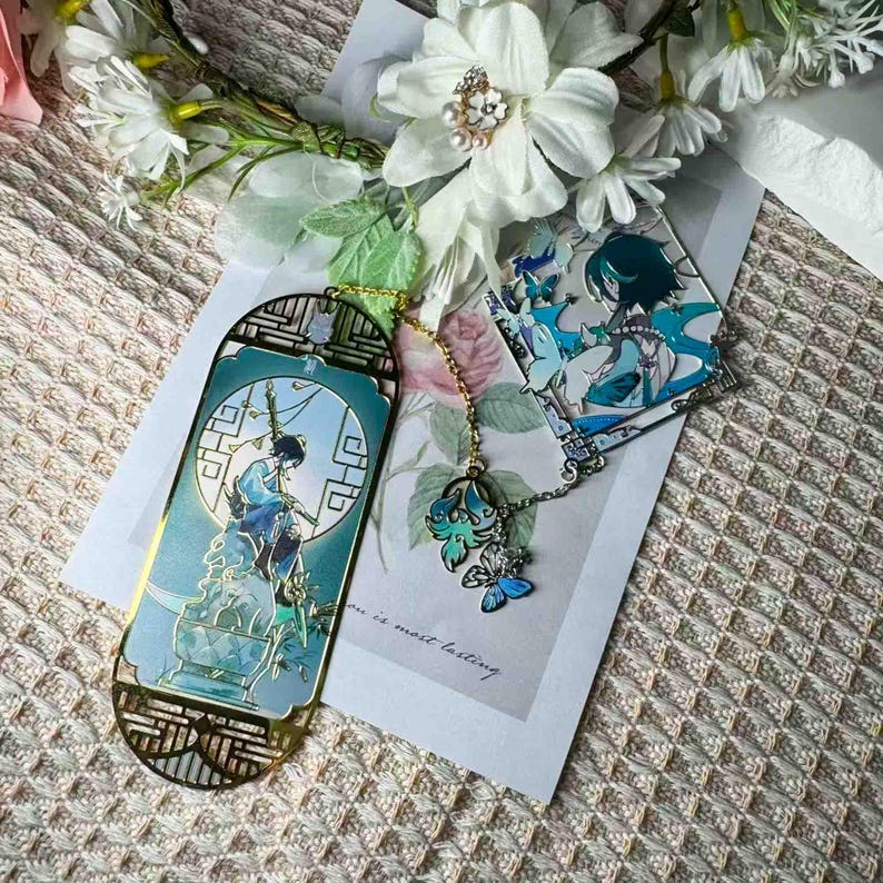 May include: A set of two metal bookmarks with a gold finish and teal and blue illustrations. One bookmark features a character illustration, and the other has a butterfly charm attached by a chain. The background includes white flowers and a card.