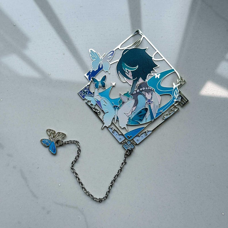 May include: A silver-toned bookmark featuring a diamond-shaped design with a character illustration and butterflies in shades of blue. A small butterfly charm is connected by a chain. The bookmark has intricate details and a decorative aesthetic.