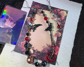 May include: A gothic-style necklace with black, red, and iridescent beads, featuring raven and gun charms, and a red teardrop pendant. The necklace is displayed on a floral background with a photo card.