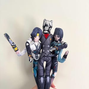 May include: Three handmade fabric dolls. The central doll has a bear-like head and wears a gray suit with a red accent. The other two dolls have dark hair and detailed outfits. The dolls are posed together, creating a group of figures.