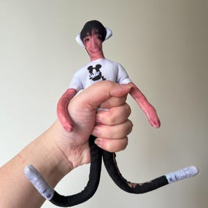 May include: A handmade doll with a printed face, wearing a white t-shirt with a black Mickey Mouse graphic. The doll has pink arms, black legs, and grey and white feet. The doll is being held in a hand.
