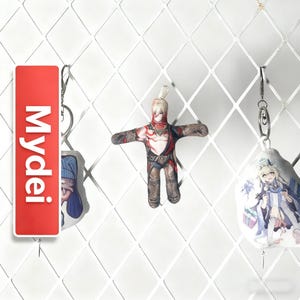 Op de afbeelding: Een verzameling sleutelhangers met anime-stijl personages en branding. E&eacute;n sleutelhanger is een rode rechthoek met het woord "Mydei" in het wit. Een andere is een kleine popachtige figuur met een rood en bruin ontwerp. De derde sleutelhanger heeft een karakterillustratie.