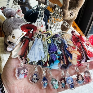 May include: A collection of anime-style acrylic keychains featuring various characters in vibrant colors. The keychains are attached to silver keyrings and are held in a hand. Some keychains depict figures in traditional Chinese attire.